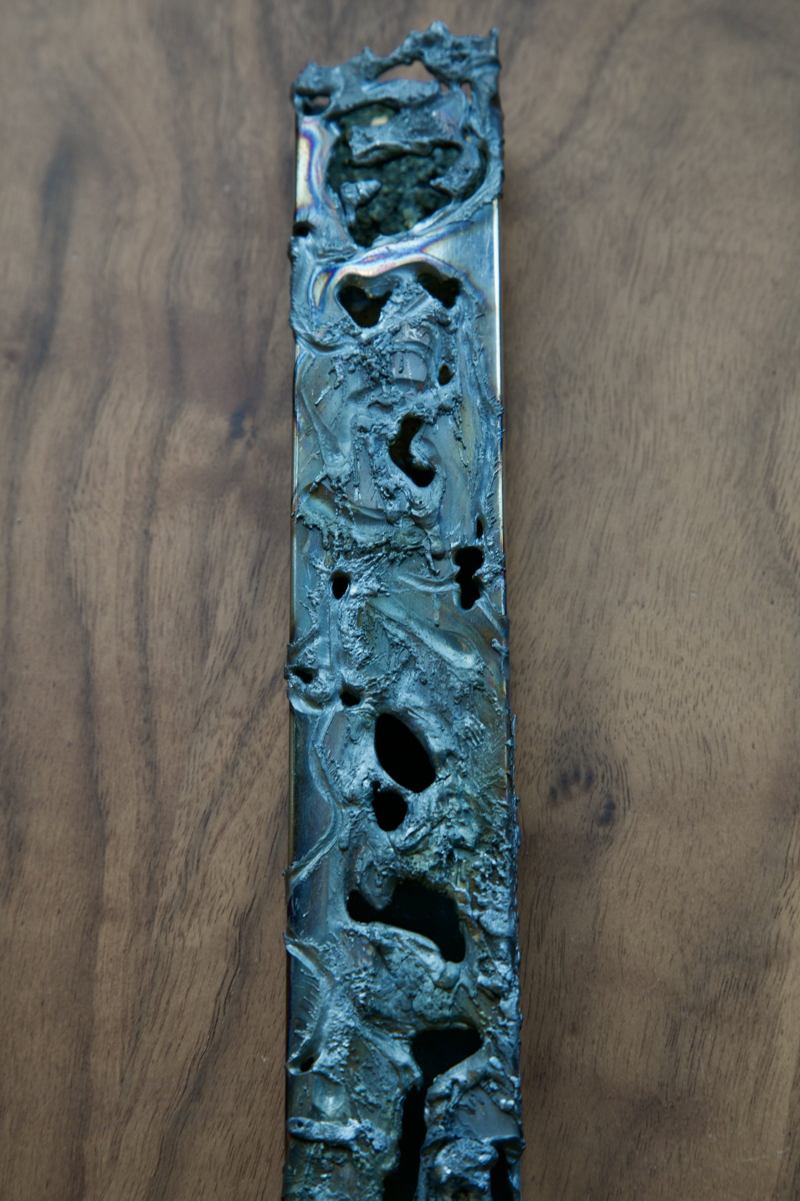 Vertical chrome steel electrode with a corroded, perforated surface, photographed against wood